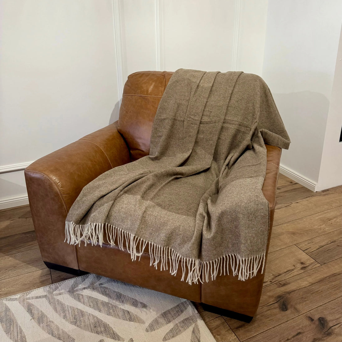 Pătură maro deschis cu franjuri albe, așezată pe fotoliu din piele